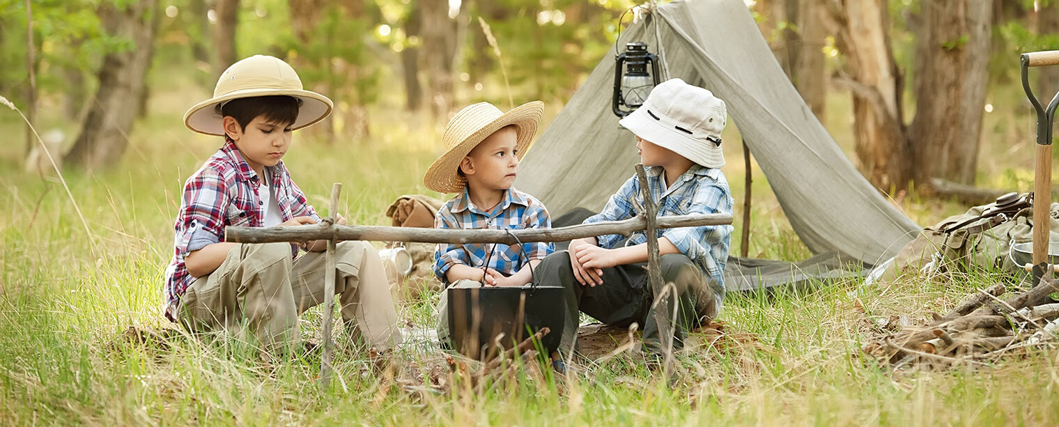 Drei Kinder sitzen im Freien um ein Lagerfeuer. Sie tragen Hüte und haben verschiedene Kleidung an. Im Hintergrund steht ein Zelt und es gibt eine grüne Waldlandschaft. Die Kinder schauen miteinander und scheinen zu plaudern.